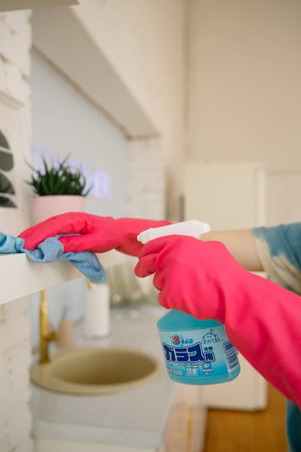 A person wearing pink rubber gloves is holding a white spray bottle with cleaning solution in one hand and a blue cloth in the other, preparing to sanitise a white kitchen shelf. The shelf, located ab