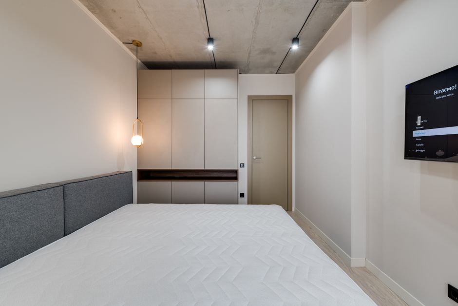 A minimalist bedroom featuring a double bed with a plain white mattress and a wooden headboard, flanked by two matching wooden bedside tables on a wooden floor. The room has white walls and built-in w