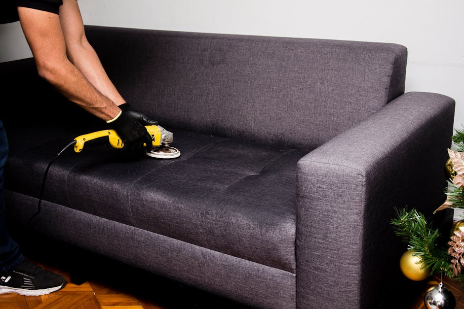 Close-up of a person's hand using a handheld vacuum cleaner to deep clean the olive-green upholstered armrest of a sofa in a living room. The sofa features textured fabric and is positioned next to a 